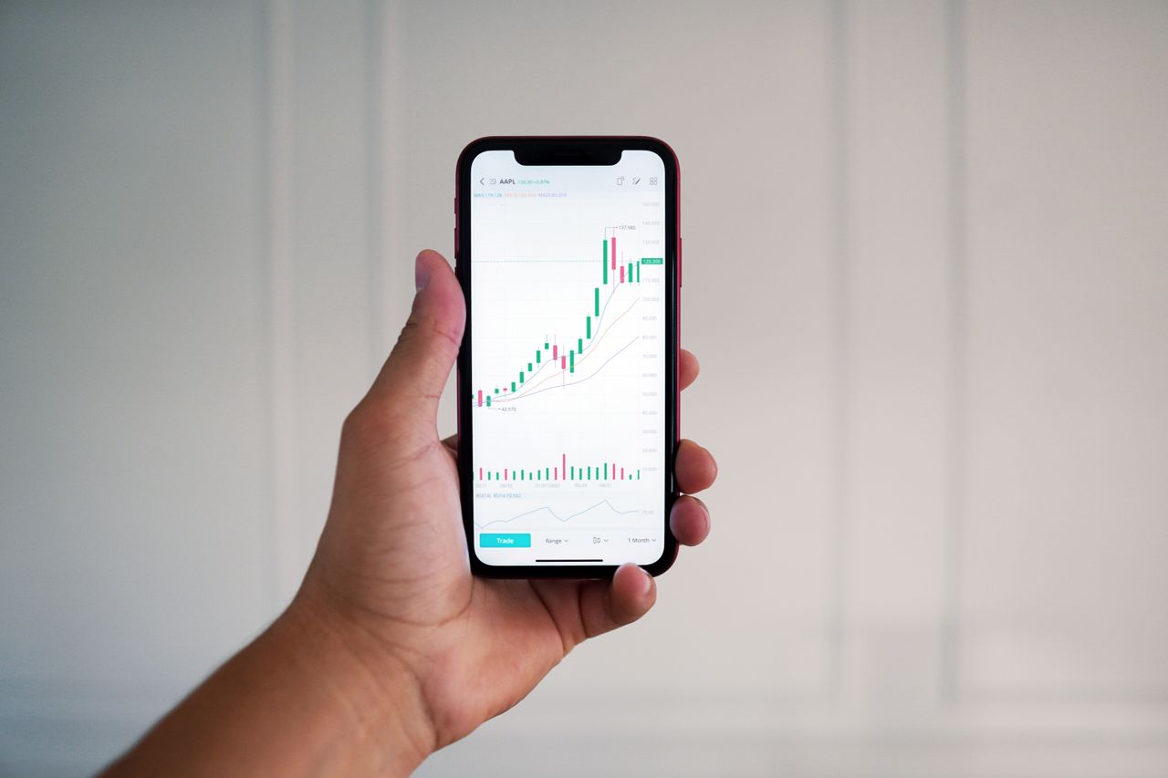 Gros plan sur une main tenant un smartphone affichant un graphique boursier en intérieur.
